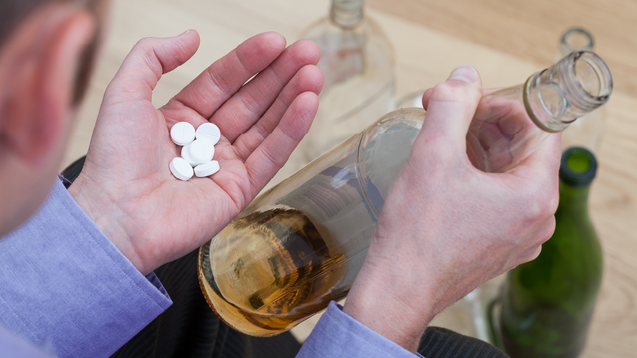 A person holding several white pills in one hand while holding a bottle of alcohol in the other, indicating a potential risk of mixing medications with alcoho