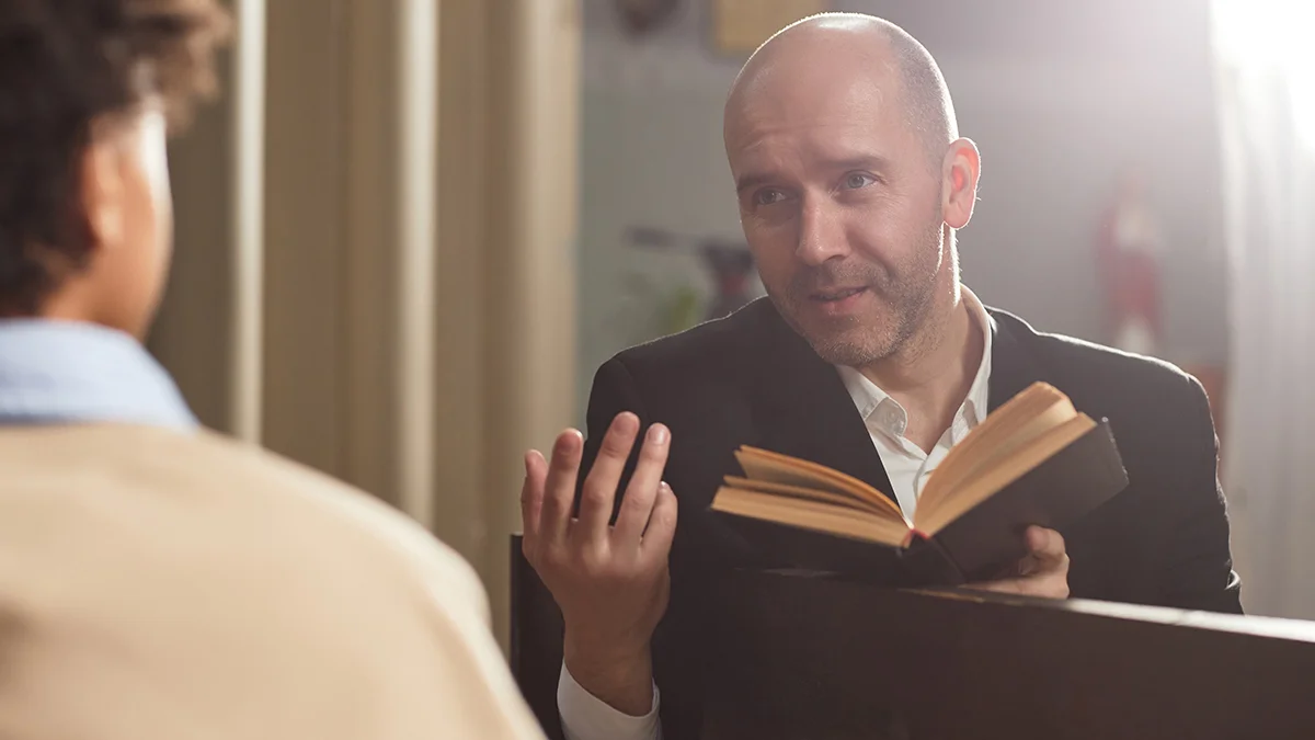 A counselor, holds an open Bible while engaging in a conversation with someone seated across from him.