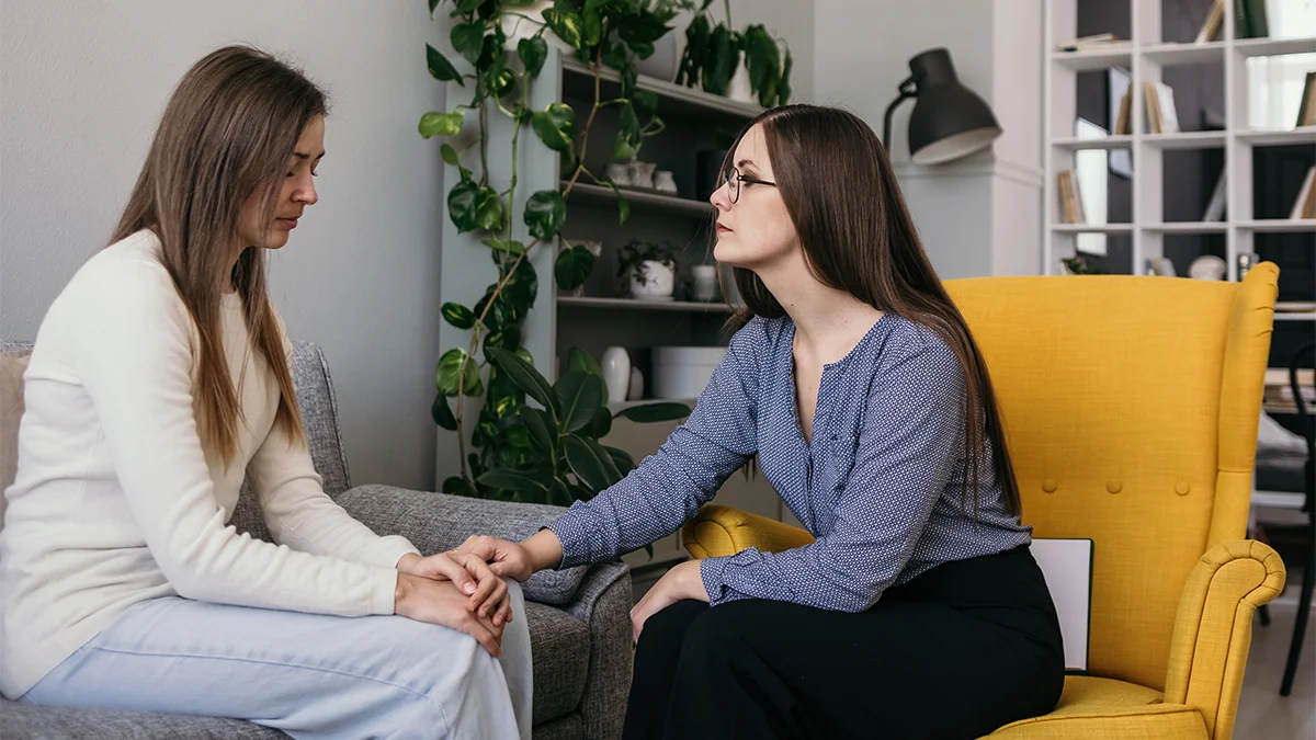 A therapist sits in a bright yellow chair, attentively holding the hands of a woman seated on a couch, offering comfort and support.