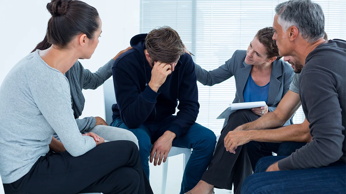 A group therapy session is taking place, with several people seated in a circle. A man in the center sits with his head in his hand, appearing distressed, while the others around him offer support by placing hands on his back and shoulders.