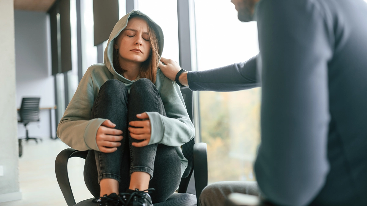 A young woman sits curled up in a chair, wearing a hoodie, with her knees pulled to her chest and her eyes closed, looking distressed or overwhelmed.