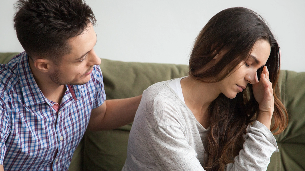 A man in a plaid shirt sits beside a woman who looks visibly upset, resting her hand on her forehead in distress.