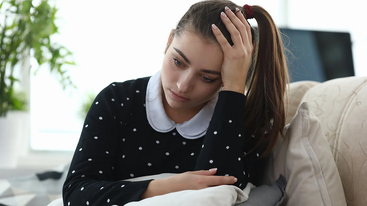 A young woman sits on a couch with her head resting on her hand, appearing sad or deep in thought.