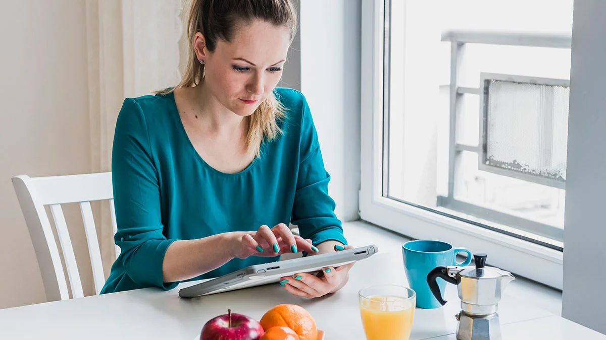 A woman sits at a table by a window, focused on using a tablet.