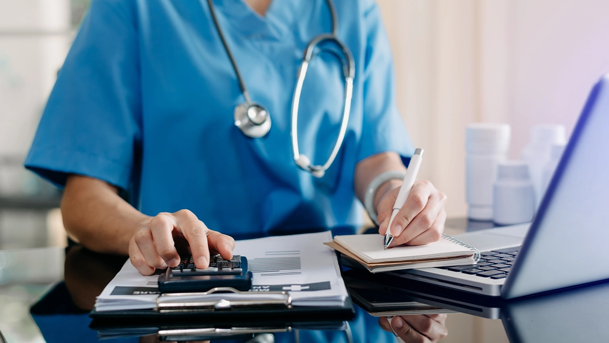 A healthcare professional wearing blue scrubs and a stethoscope works at a desk, using a calculator while writing notes in a notebook.