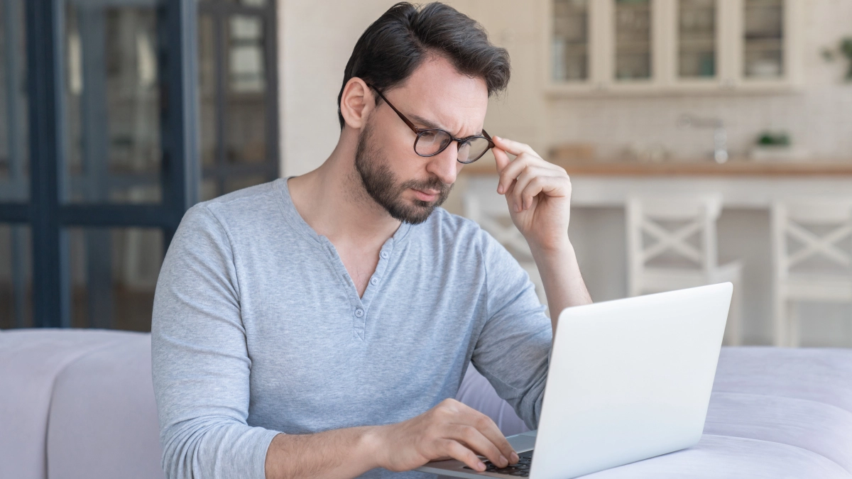 A man sits at a desk working on a laptop, adjusting his glasses and looking at the screen with a focused and slightly concerned expression.