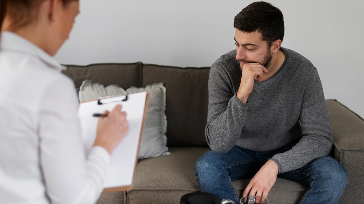 A man sits on a couch, appearing deep in thought during a counseling session. He rests his chin on his hand and looks contemplative. In the foreground, a healthcare professional holds a clipboard, taking notes while speaking with him.