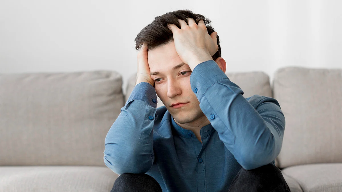 A young man sits on a couch with his hands resting on his head, looking frustrated or overwhelmed.