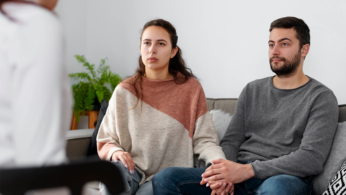 A couple sits on a couch, holding hands during a therapy or counseling session.