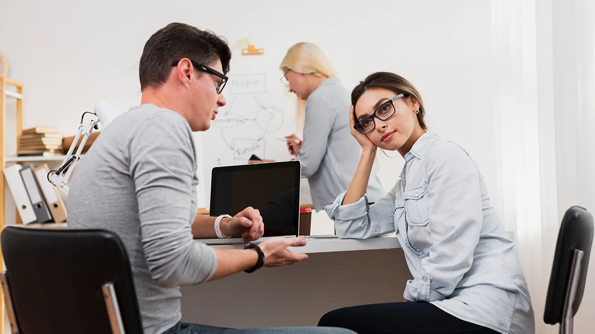 A man and woman are seated at a desk, engaged in a conversation. The man gestures with his hands, explaining something, while the woman rests her head on her hand, looking thoughtful or slightly frustrated.