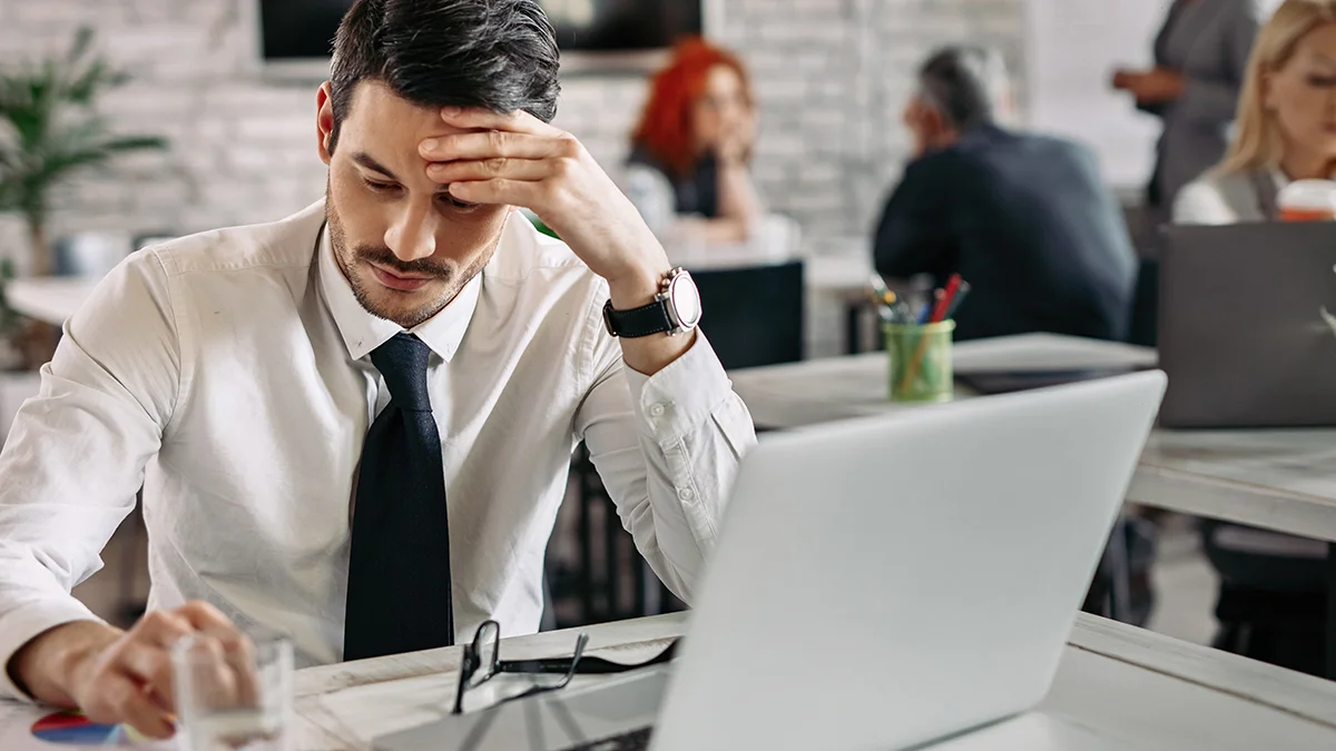 A man in a white shirt and tie sits at a desk, looking stressed as he rests his forehead on his hand.