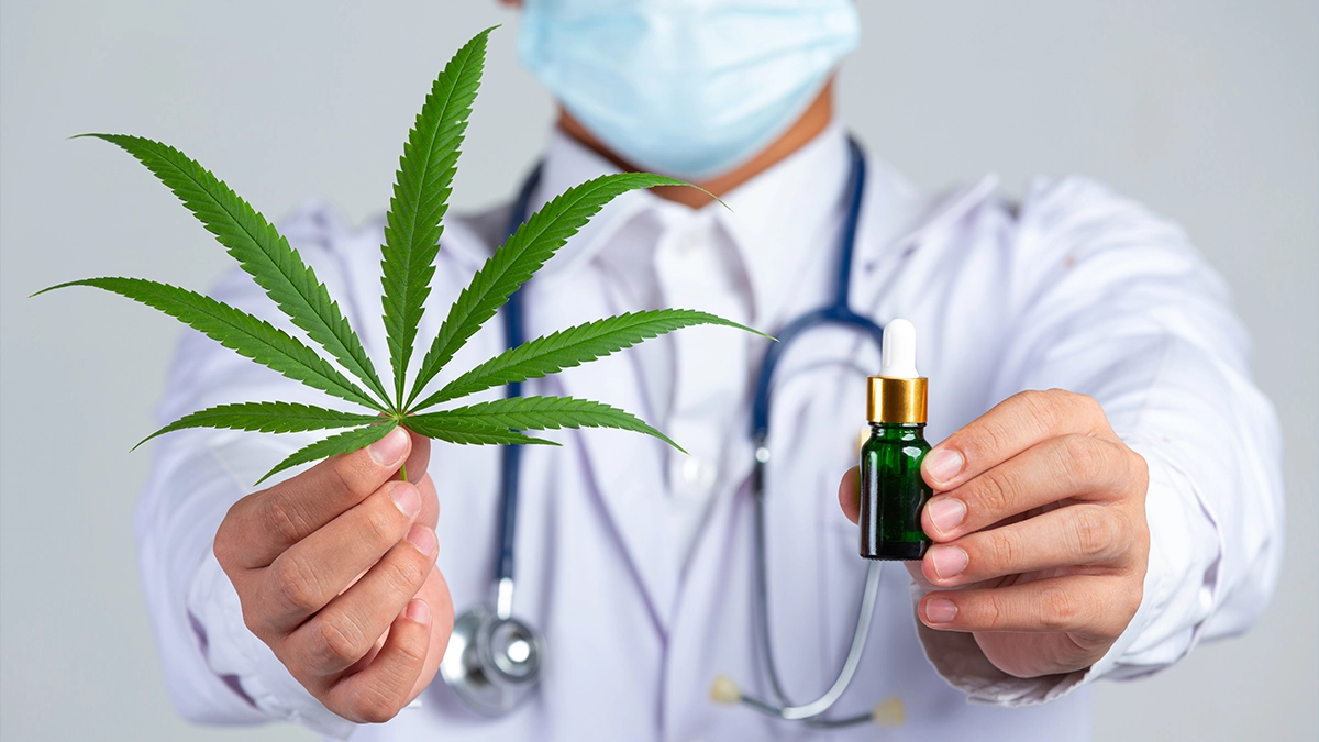 A doctor wearing a white coat, stethoscope, and a medical mask holds up a cannabis leaf in one hand and a small green bottle with a dropper in the other.